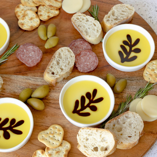 Wooden board with bread, olives, salami, and bowls of yellow dip.