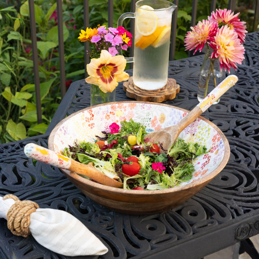 Bowl of salad with a wooden spoon on a garden table with flowers and lemonade.