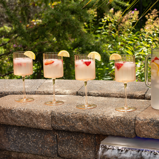 Four glasses of pink cocktails with lemon slices on a stone ledge outdoors.