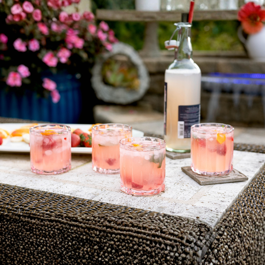 Pink cocktails with ice in glasses on a stone table outdoors, with flowers and a bottle in the background.