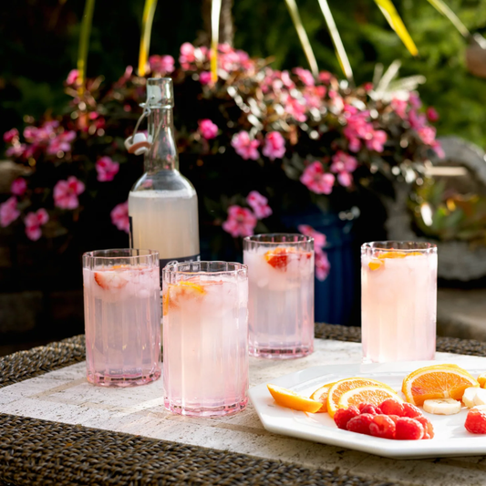 Outdoor setting with pink cocktails in glasses and a bottle, surrounded by flowers and fruit.