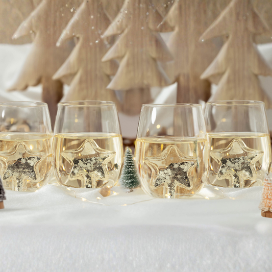 Four glasses of champagne with decorative stars in a snowy scene with Christmas trees.