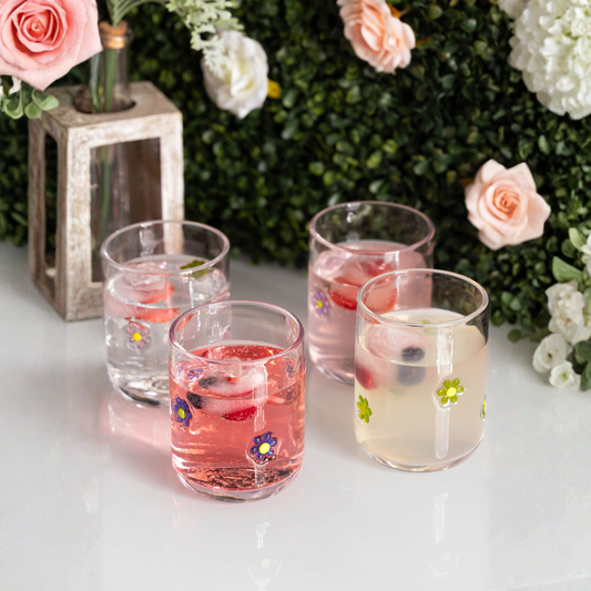 Four glasses of pink and clear drinks with floral decorations on a white surface with flowers and lantern in the background.