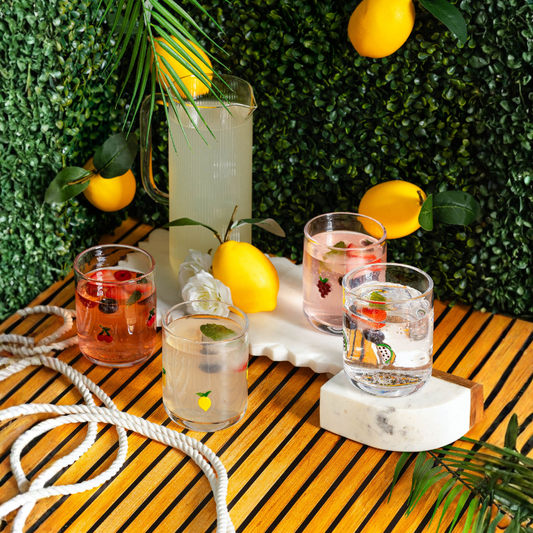 Outdoor setting with lemonade pitcher and glasses on a wooden table with lemons and greenery.