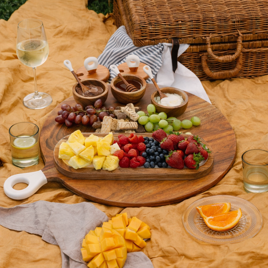 Picnic setup with a wooden platter of fruits, cheese, and honey, accompanied by glasses of wine and juice on a yellow blanket.