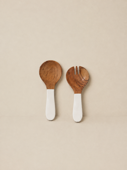 Two wooden utensils with white handles, a spoon on the left and a small fork on the right, displayed on a neutral beige background.
