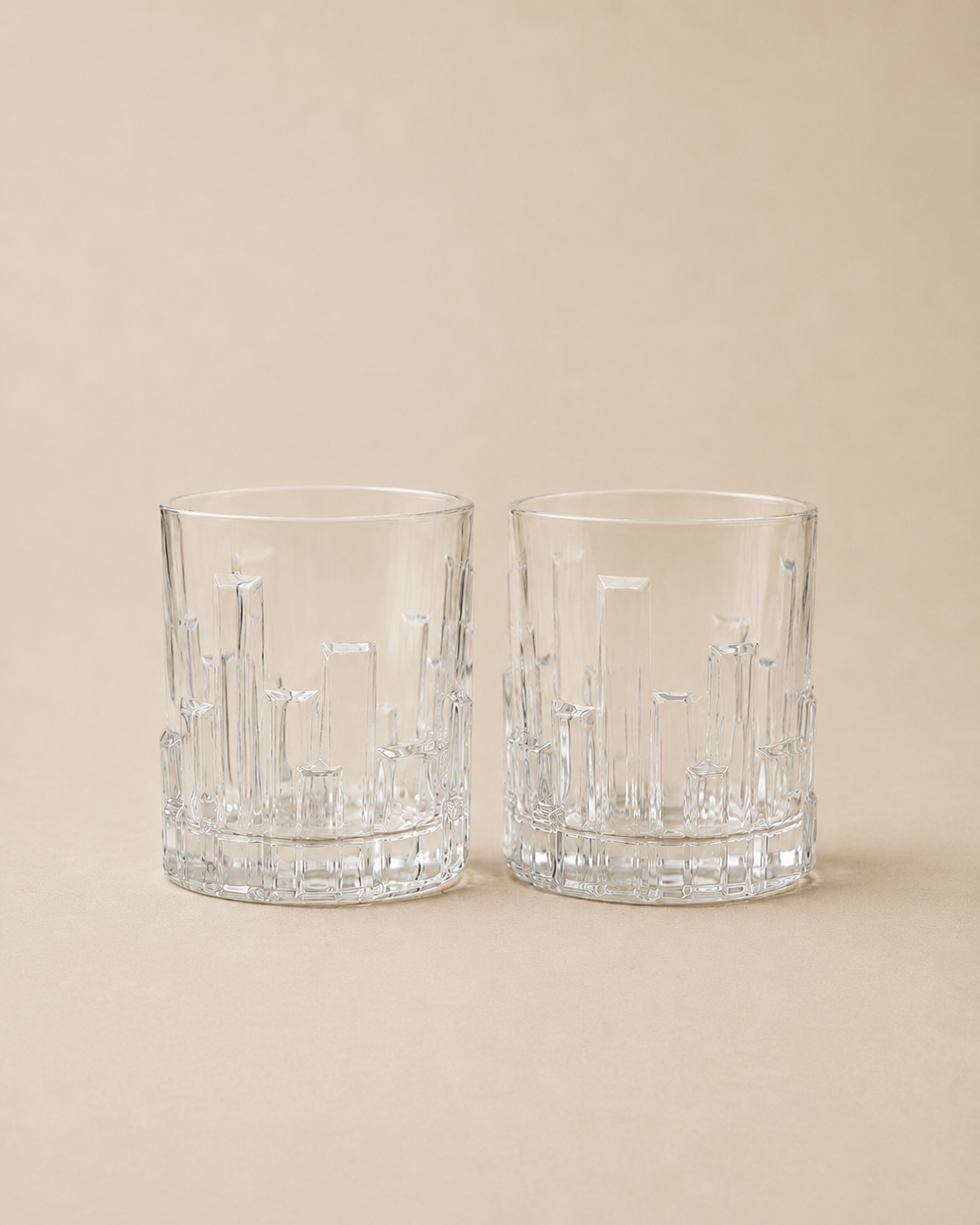 Two clear textured whiskey glasses with a geometric, faceted design, set against a solid beige background.
