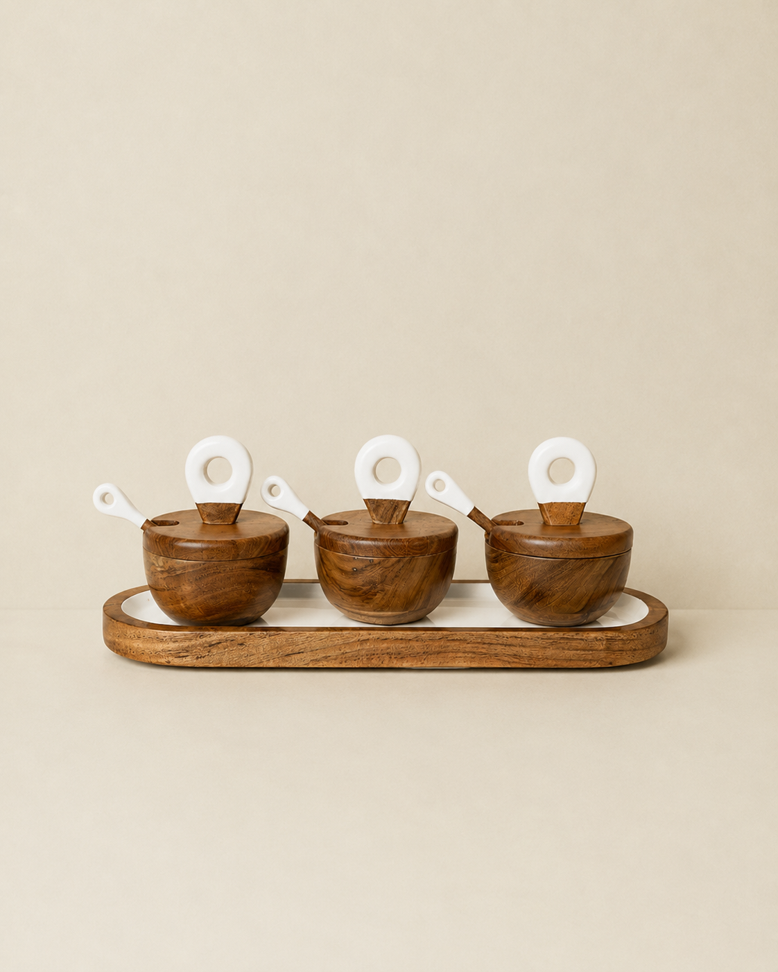 Three small wooden bowls with matching lids and white spoon handles arranged on a long wooden tray against a plain beige background.