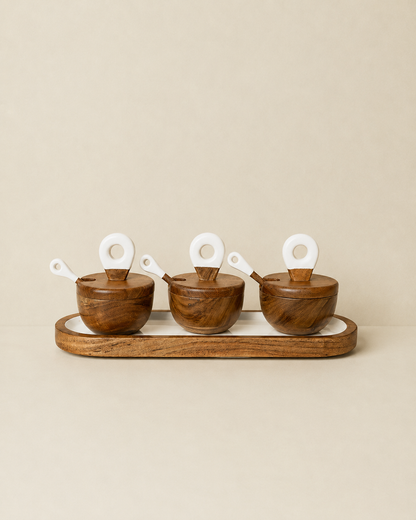 Three small wooden bowls with matching lids and white spoon handles arranged on a long wooden tray against a plain beige background.
