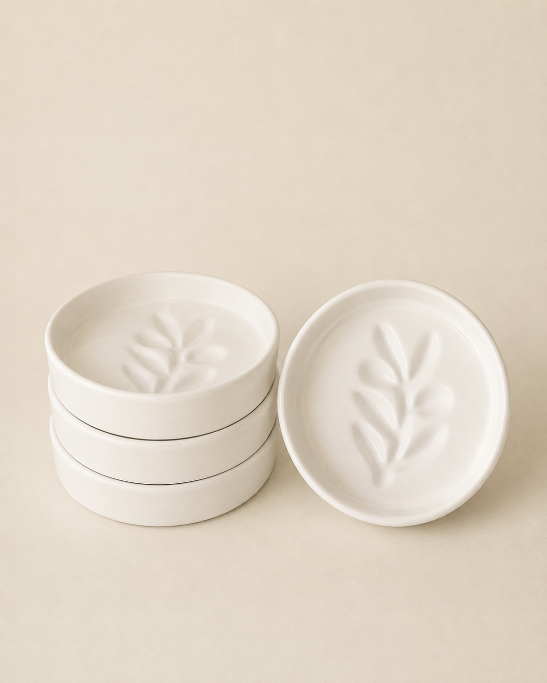 Three small round white ceramic dishes with embossed leaf motifs, with one dish standing open to the right and the other two stacked on the left, against a warm beige background.