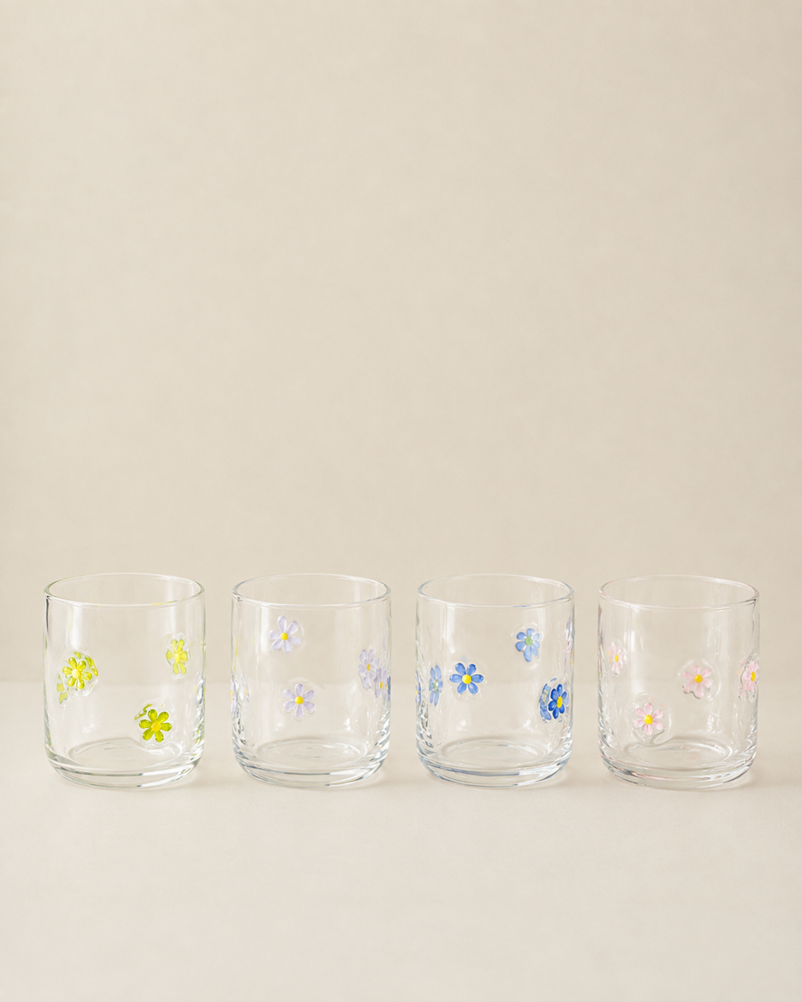 Four clear glass tumblers with small hand-painted floral designs in yellow, blue, pink, and white, lined up on a light beige background.