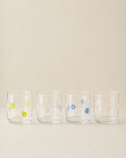 Four clear glass tumblers with small hand-painted floral designs in yellow, blue, pink, and white, lined up on a light beige background.