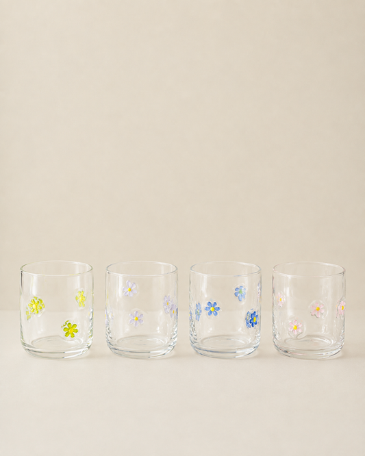 Four clear glass tumblers with small hand-painted floral designs in yellow, blue, pink, and white, lined up on a light beige background.