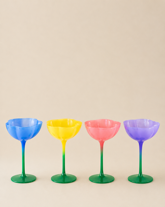 Four flower-shaped cocktail glasses lined up in a row, featuring blue, yellow, pink, and purple gradient stems and bases on a solid beige background.