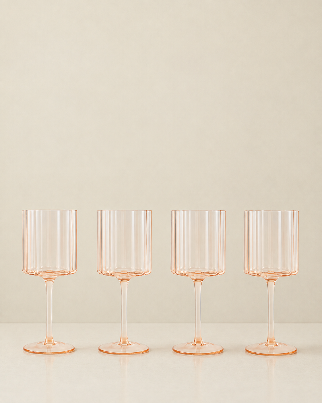 Four clear stemware glasses with a delicate peach-pink tint, arranged in a row on a light surface against a warm beige background.