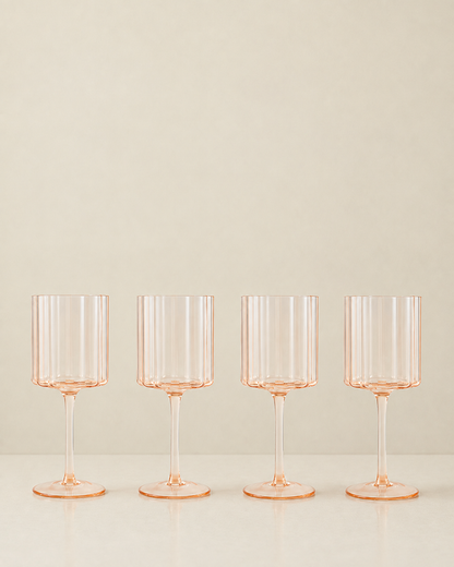 Four clear stemware glasses with a delicate peach-pink tint, arranged in a row on a light surface against a warm beige background.