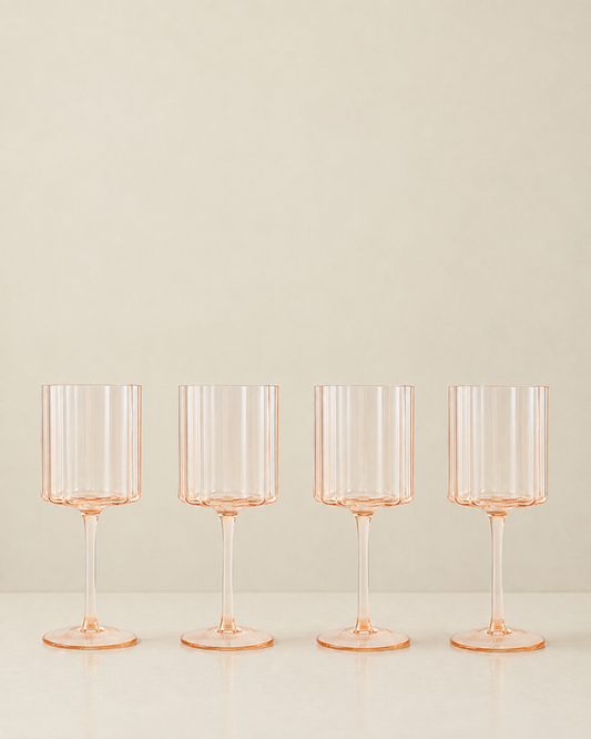 Four clear stemware glasses with a delicate peach-pink tint, arranged in a row on a light surface against a warm beige background.