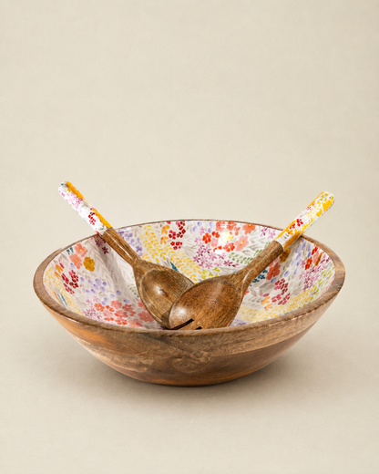 A rustic wooden salad bowl with a colorful floral pattern on the interior, accompanied by two wooden spoons with patterned handles, set against a solid beige background.