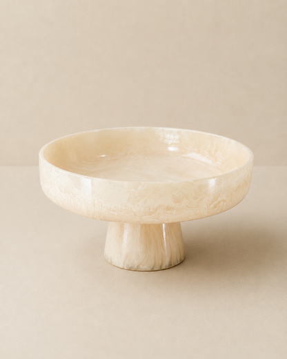 A round pedestal bowl made of cream-colored carved wood with a natural marbled texture, set against a matching beige background.