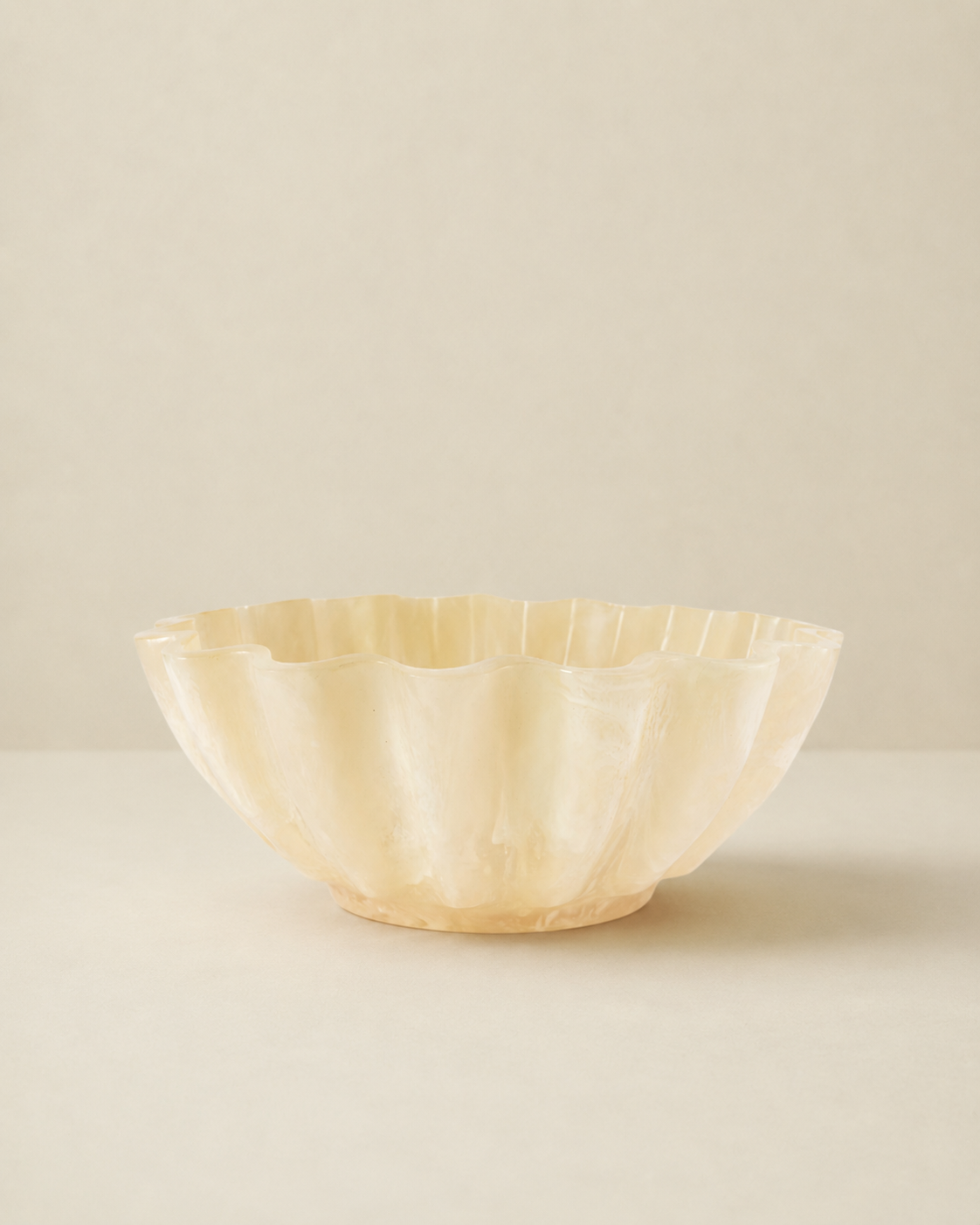 A scalloped bowl made of a warm, creamy ivory material, likely bone or mother-of-pearl, set against a minimalist beige background.