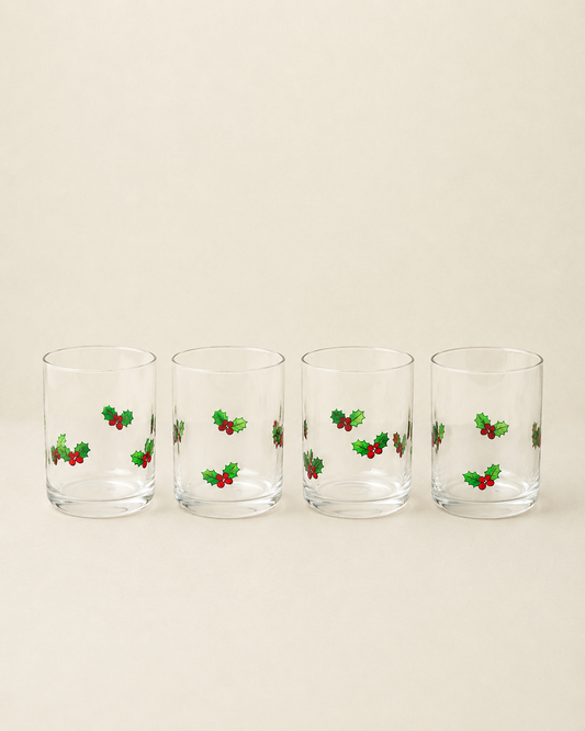 Four clear lowball glasses with small green holly and red berry motifs, arranged in a row on a warm beige background.