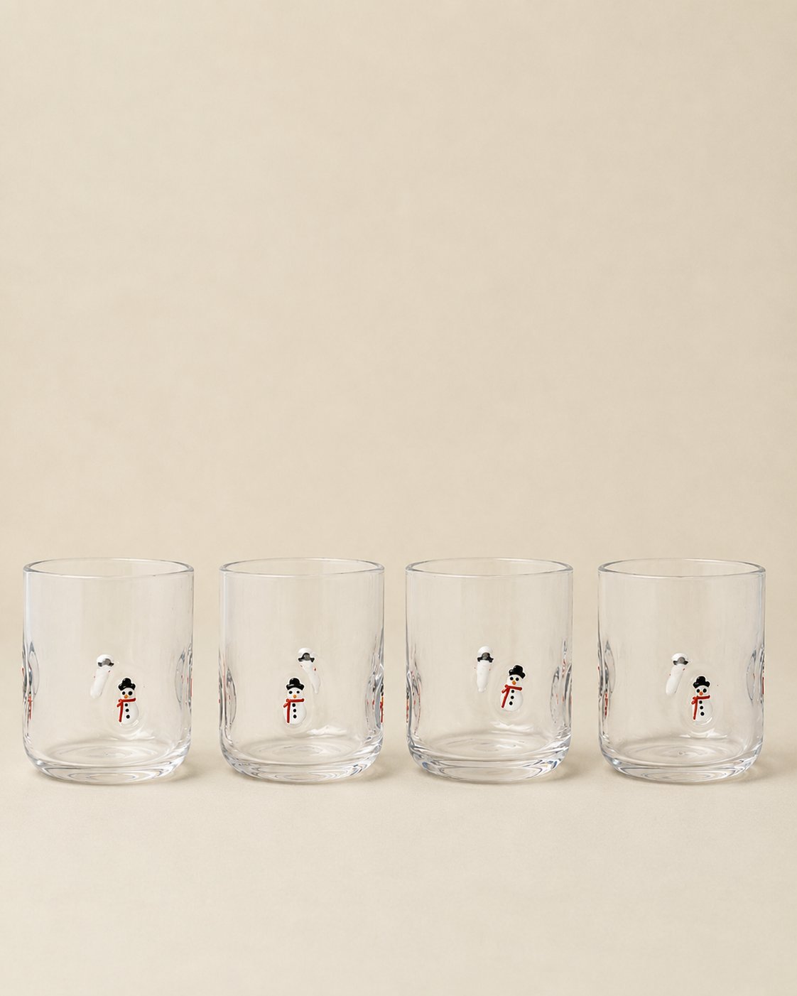 Four clear glass tumblers with small illustrated snowman designs, arranged in a row against a warm beige background.