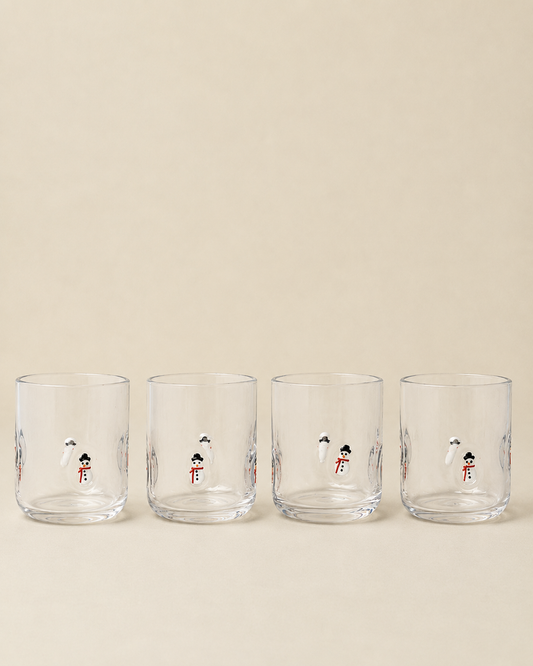Four clear glass tumblers with small illustrated snowman designs, arranged in a row against a warm beige background.