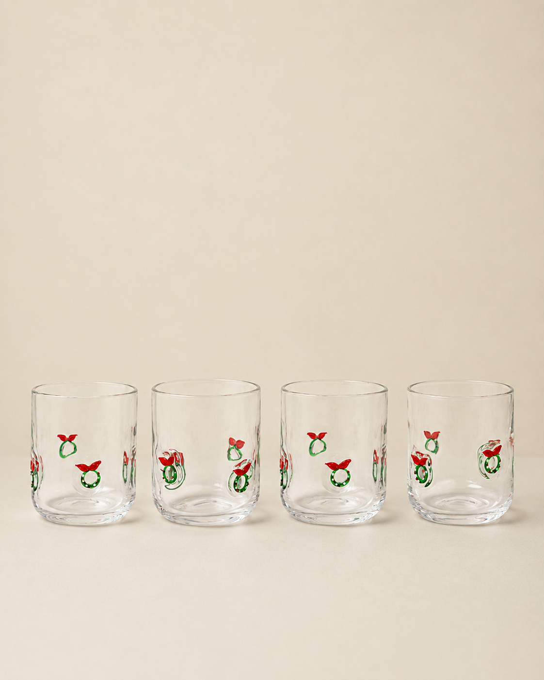 Four clear glass tumblers with a repeating Christmas wreath and red bow pattern, arranged in a row against a solid beige background.