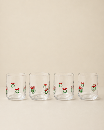 Four clear glass tumblers with a repeating Christmas wreath and red bow pattern, arranged in a row against a solid beige background.