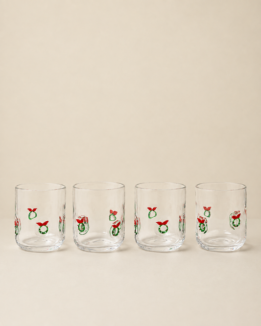 Four clear glass tumblers with a repeating Christmas wreath and red bow pattern, arranged in a row against a solid beige background.