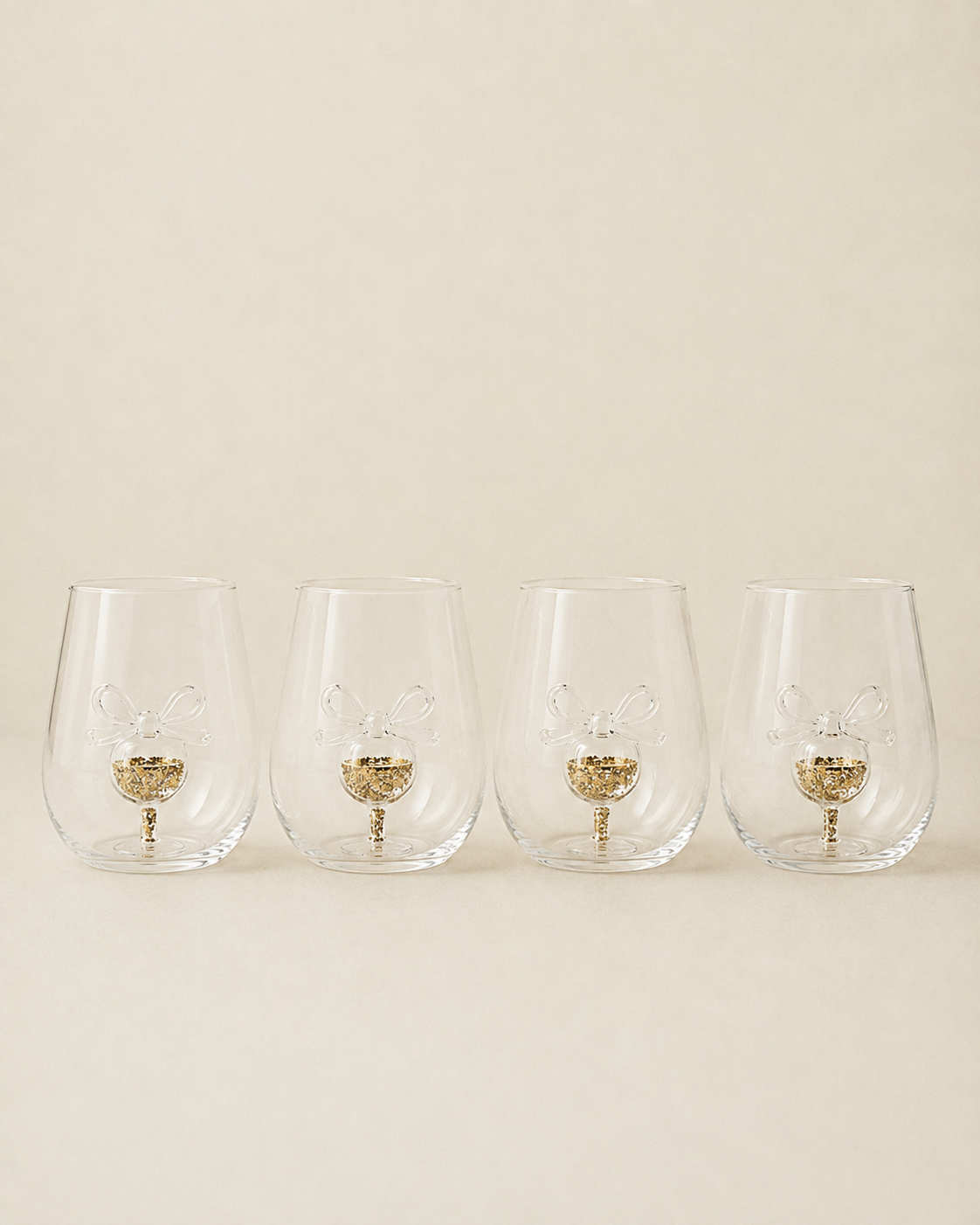 Four clear stemless wine glasses with a gold glitter bunny design in the base, arranged in a row against a solid beige background.