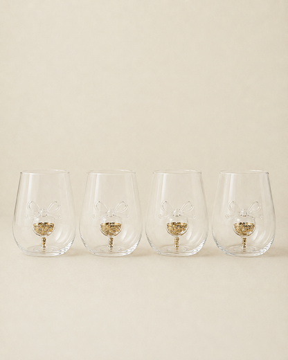 Four clear stemless wine glasses with a gold glitter bunny design in the base, arranged in a row against a solid beige background.