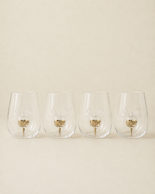 Four clear stemless wine glasses with a gold glitter bunny design in the base, arranged in a row against a solid beige background.