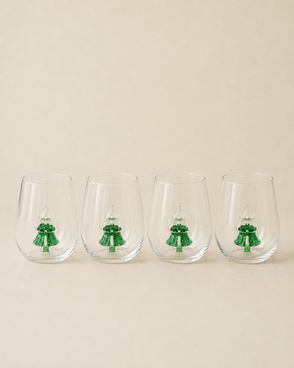 Four clear stemless wine glasses lined up in a row, each decorated with a small green Christmas tree design near the base, set against a solid beige background.
