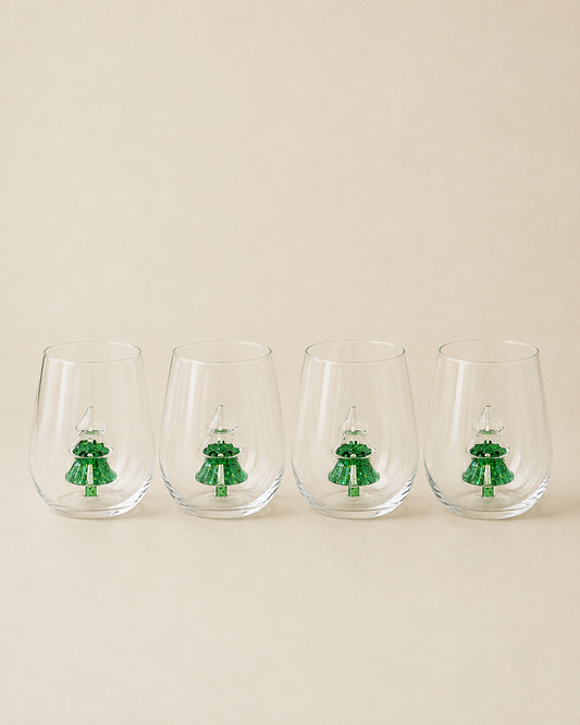 Four clear stemless wine glasses lined up in a row, each decorated with a small green Christmas tree design near the base, set against a solid beige background.