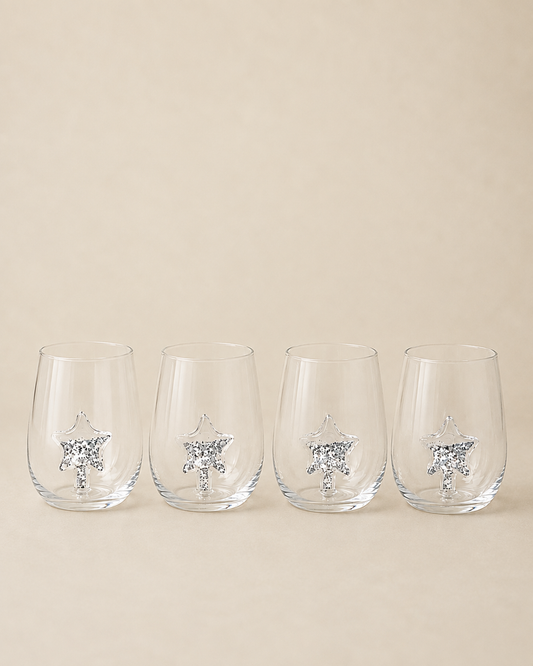 Four clear stemless wine glasses with a silver glittery star design near the base, arranged in a row against a solid beige background.