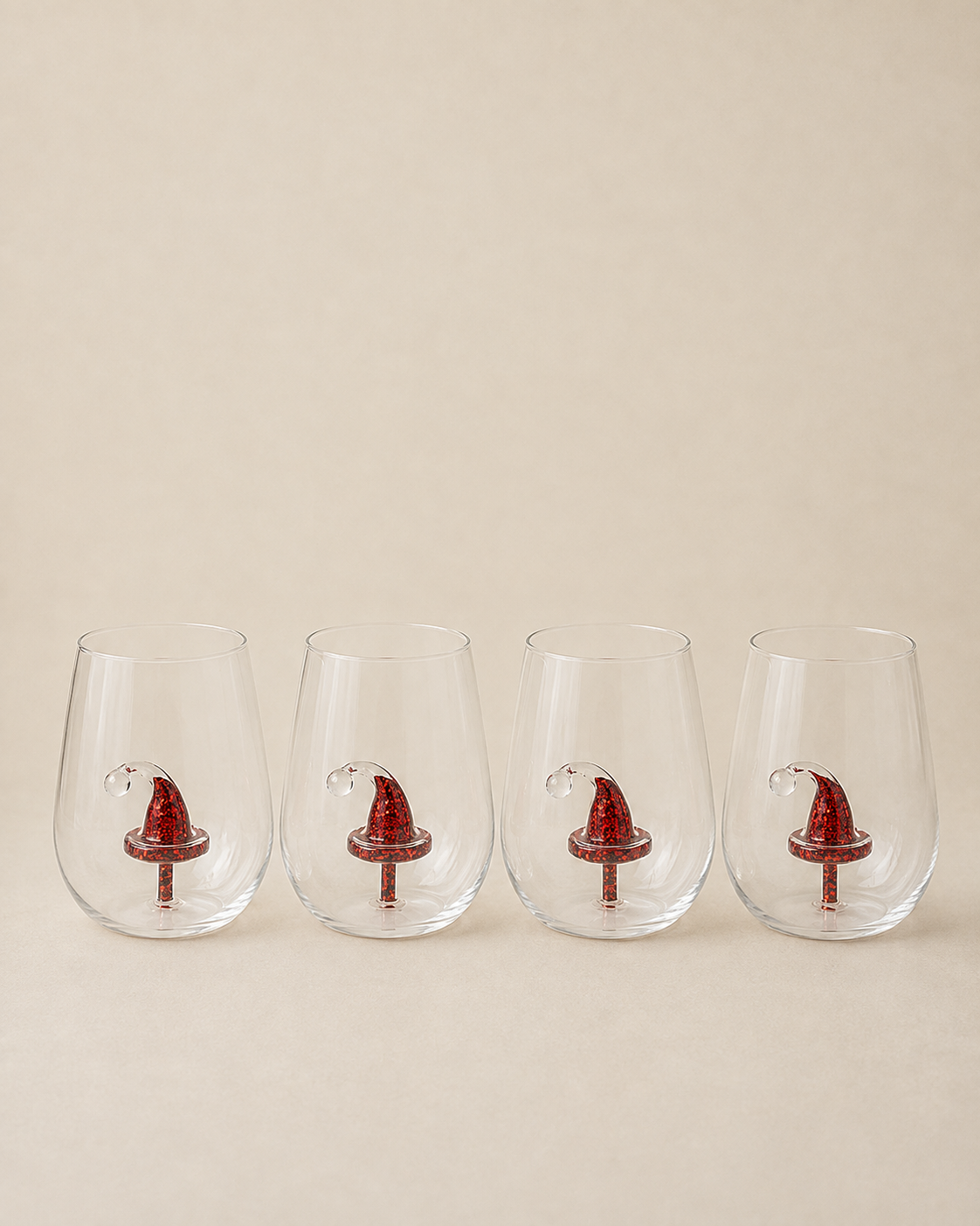 Four clear stemless wine glasses lined up in a row, each decorated with a small red Santa hat near the base, set against a solid beige background.