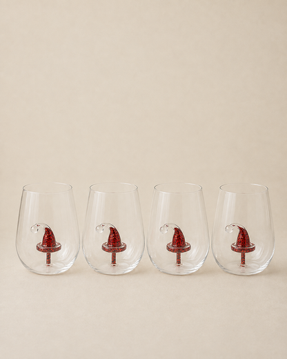 Four clear stemless wine glasses lined up in a row, each decorated with a small red Santa hat near the base, set against a solid beige background.
