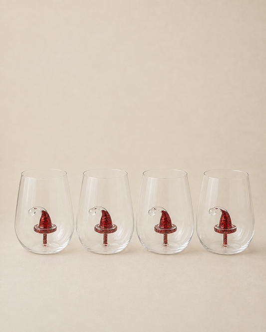 Four clear stemless wine glasses lined up in a row, each decorated with a small red Santa hat near the base, set against a solid beige background.