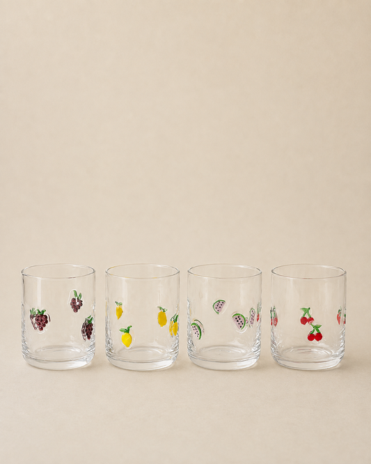 Four clear glass tumblers with small colorful fruit illustrations, arranged in a row on a warm beige background.