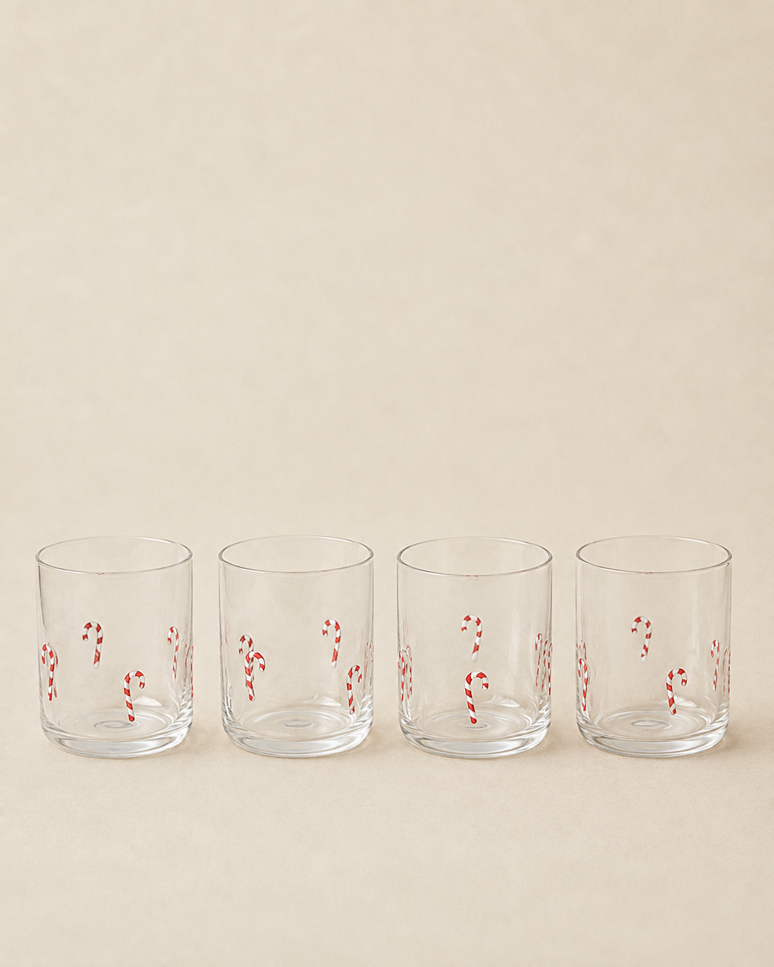 Four clear lowball glasses with red and white candy cane patterns, lined up in a row against a solid beige background.
