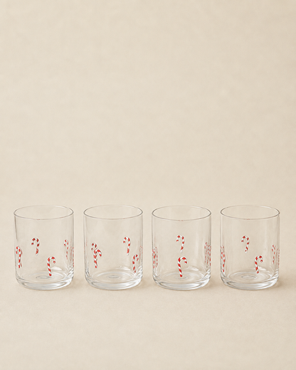 Four clear lowball glasses with red and white candy cane patterns, lined up in a row against a solid beige background.