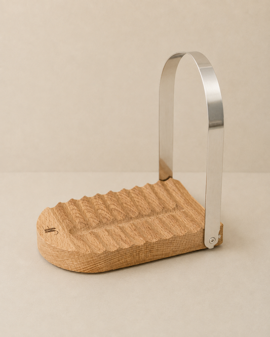 A minimalist wooden footrest with a wavy textured top surface and a curved, polished silver metal support arm, set against a neutral beige background.